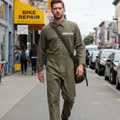 Man in green jumpsuit walking on a city street with a bike repair shop in the background.