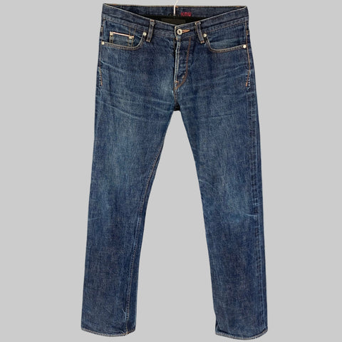A pair of KICKING MULE WORKSHOP indigo cotton button fly jeans displayed on a hanger, showing a straight-leg cut and button fly closure.